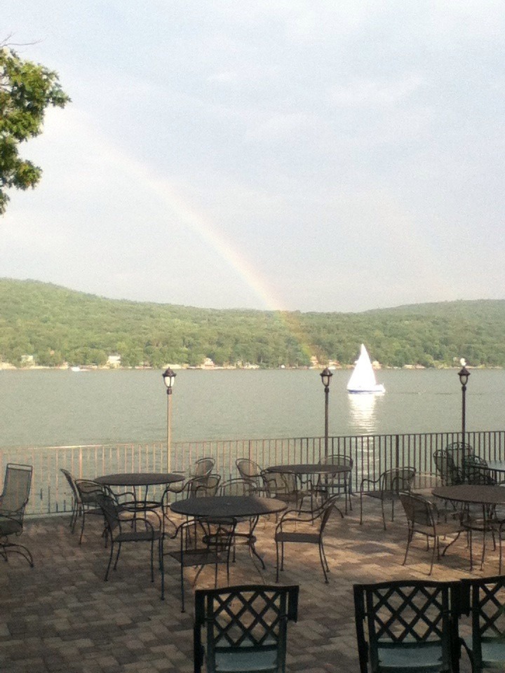 Sailboat glides across a lake under a rainbow, with empty patio tables in the foreground, capturing a serene, picturesque landscape.