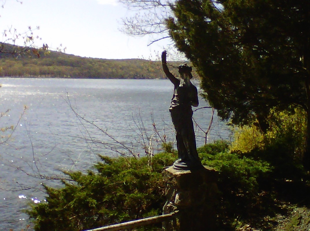 A statue resembling a woman standing on a rock near a river, with raised arm and flowing garments, surrounded by trees and distant hills.