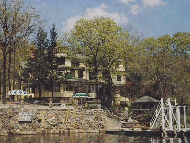 Historic lakeside lodge with a stone patio and green trees, featuring a dock and a sign reading "Continental." A peaceful retreat scene.