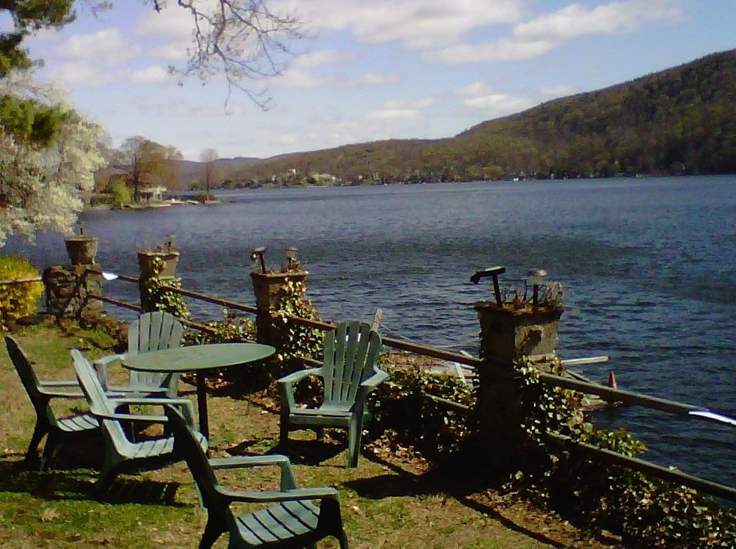 Lakeside seating area with green chairs and a table overlooks a serene blue lake, surrounded by hills and trees under a partly cloudy sky.