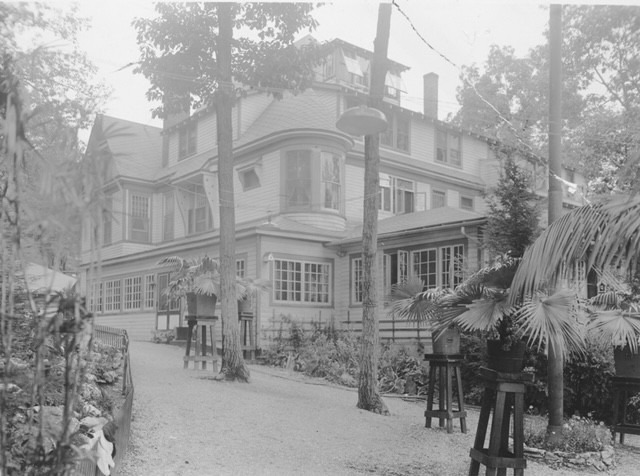 Victorian-style house surrounded by trees, with a gravel path leading to the entrance. Potted plants line the walkway, suggesting a serene garden setting.