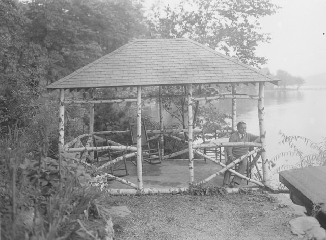 A man sits on a log near a rustic gazebo by a lake, surrounded by greenery. The image captures a serene outdoor setting.