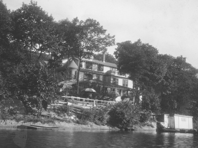 Lakefront house surrounded by trees, featuring balconies and umbrellas. A boat dock is visible in the foreground, suggesting recreational use.
