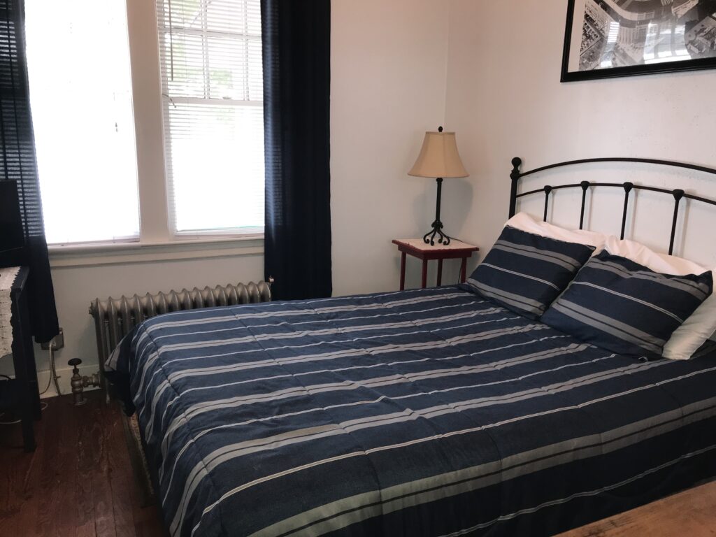 Cozy bedroom featuring a striped blue comforter, two pillows, a small lamp, and large windows with black curtains, creating a serene atmosphere.