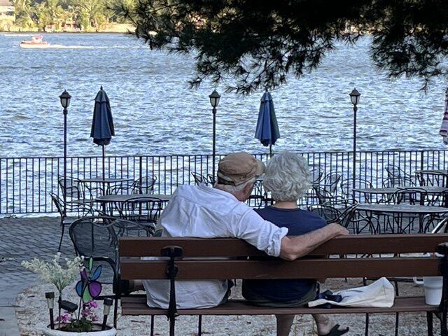 An elderly couple sits closely on a bench, overlooking a serene lake. Blue umbrellas and empty tables create a tranquil waterfront scene.