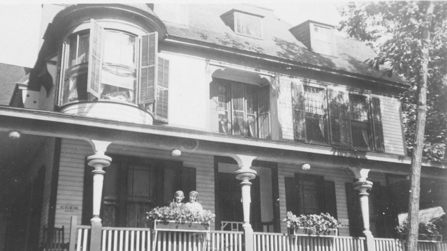 Two children stand on the porch of a vintage house with a curved bay window, surrounded by lush greenery and flower boxes.