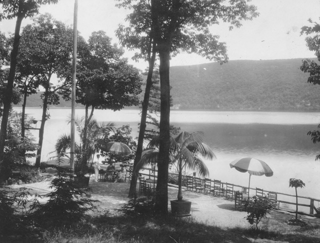 Lakeside scene featuring trees, umbrellas, and a dock reflecting a peaceful nature setting. This historic photo highlights leisure and outdoor beauty.