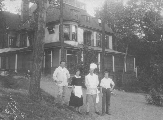 Staff stand in front of a large, historic house, dressed in early 20th-century clothing. The image highlights the setting's social context and service history.