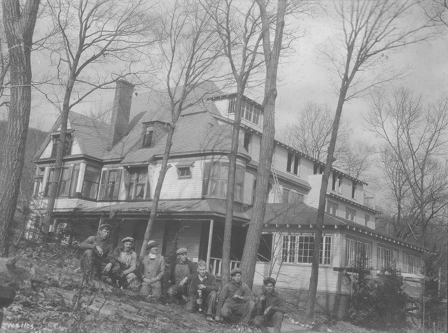 Group of men seated on a hillside in front of a large, old house surrounded by bare trees, showcasing early 20th-century architecture.